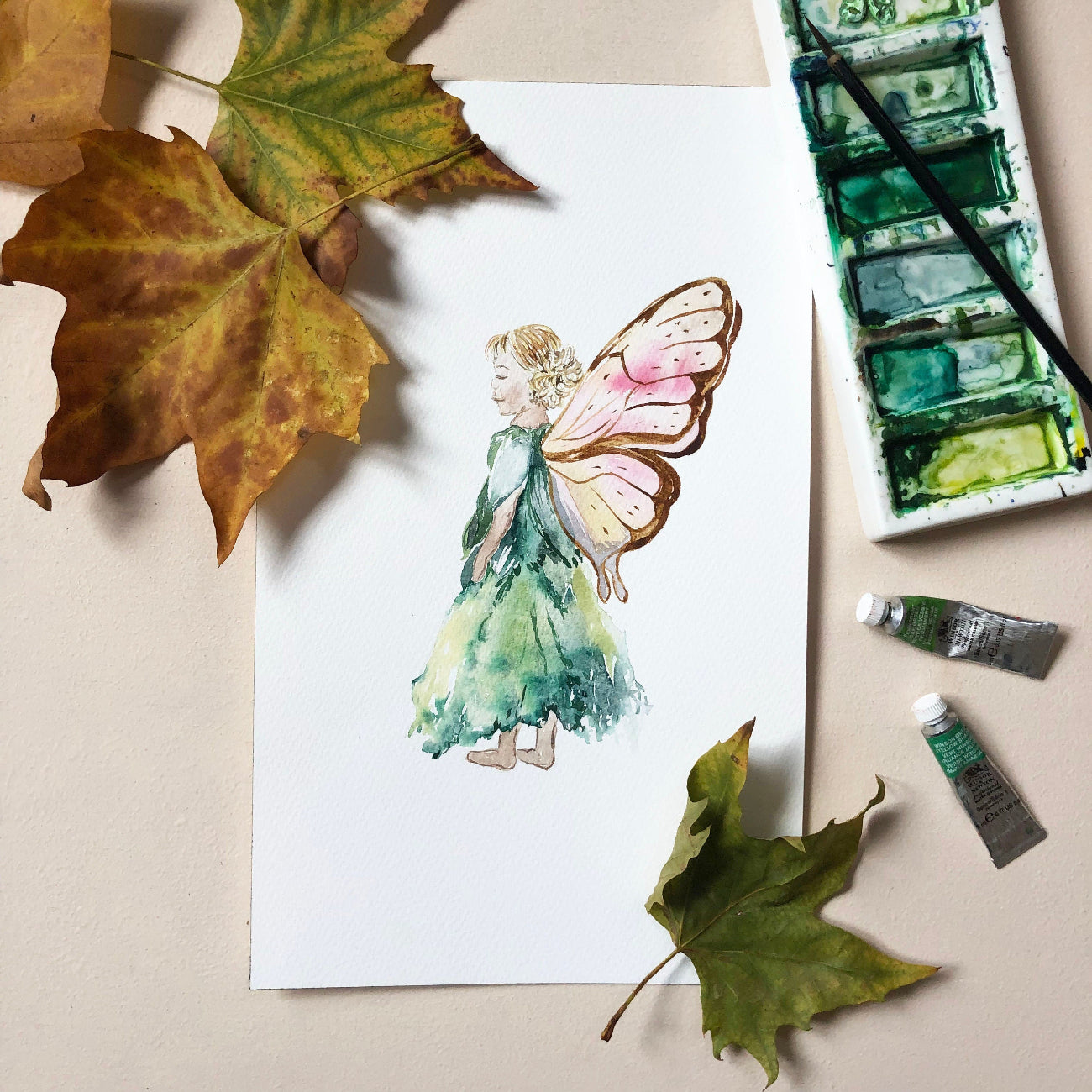 Watercolor painting of a fairy on paper with leaves and art supplies.