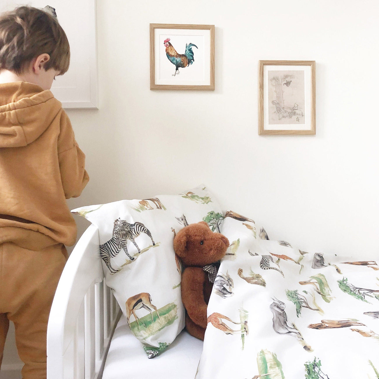 Child in a brown outfit standing next to a crib with animal-patterned bedding, framed artwork on the wall.
